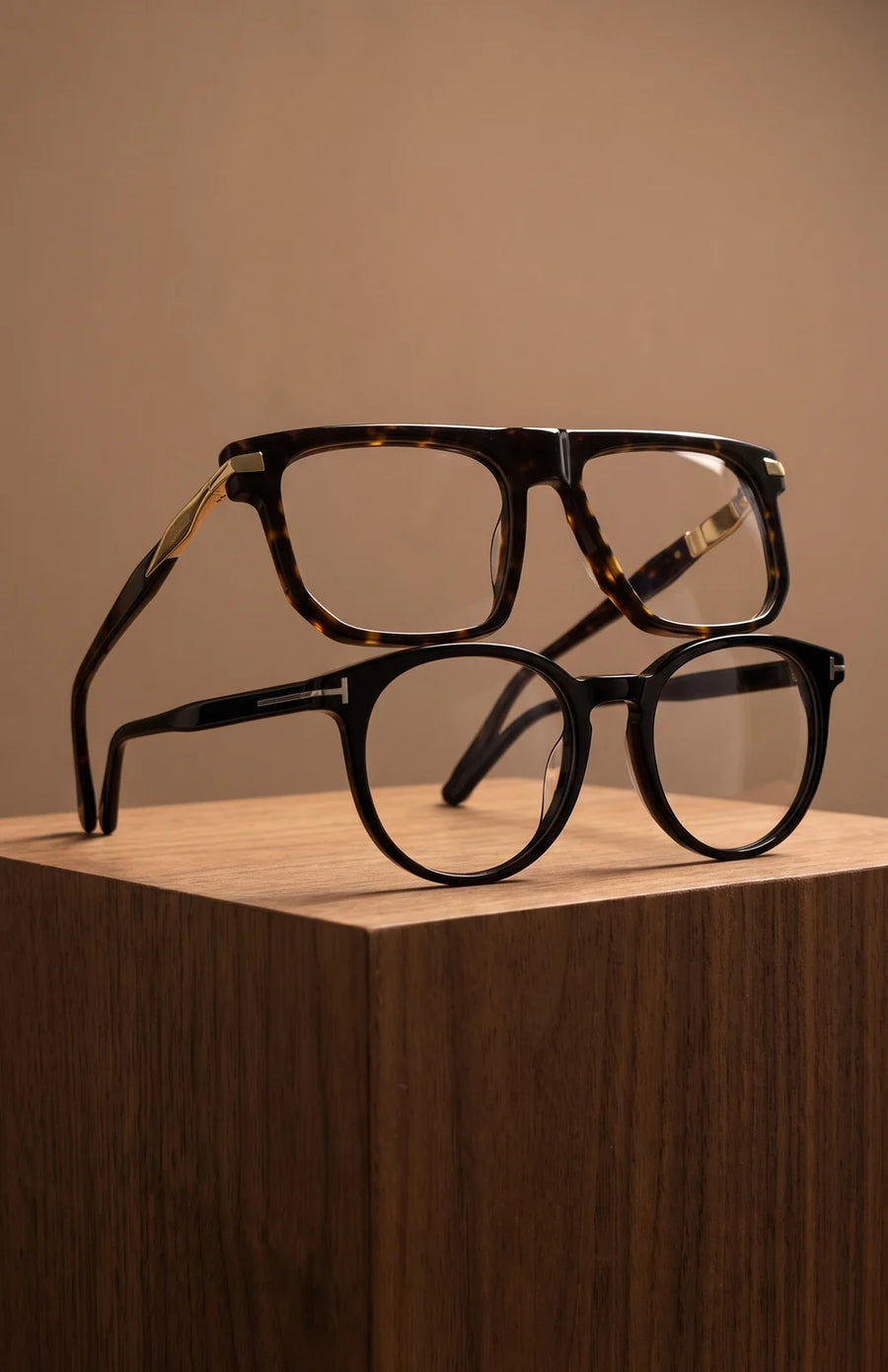 Two acetate eyeglasses, tortoise and black, stacked on a walnut block.