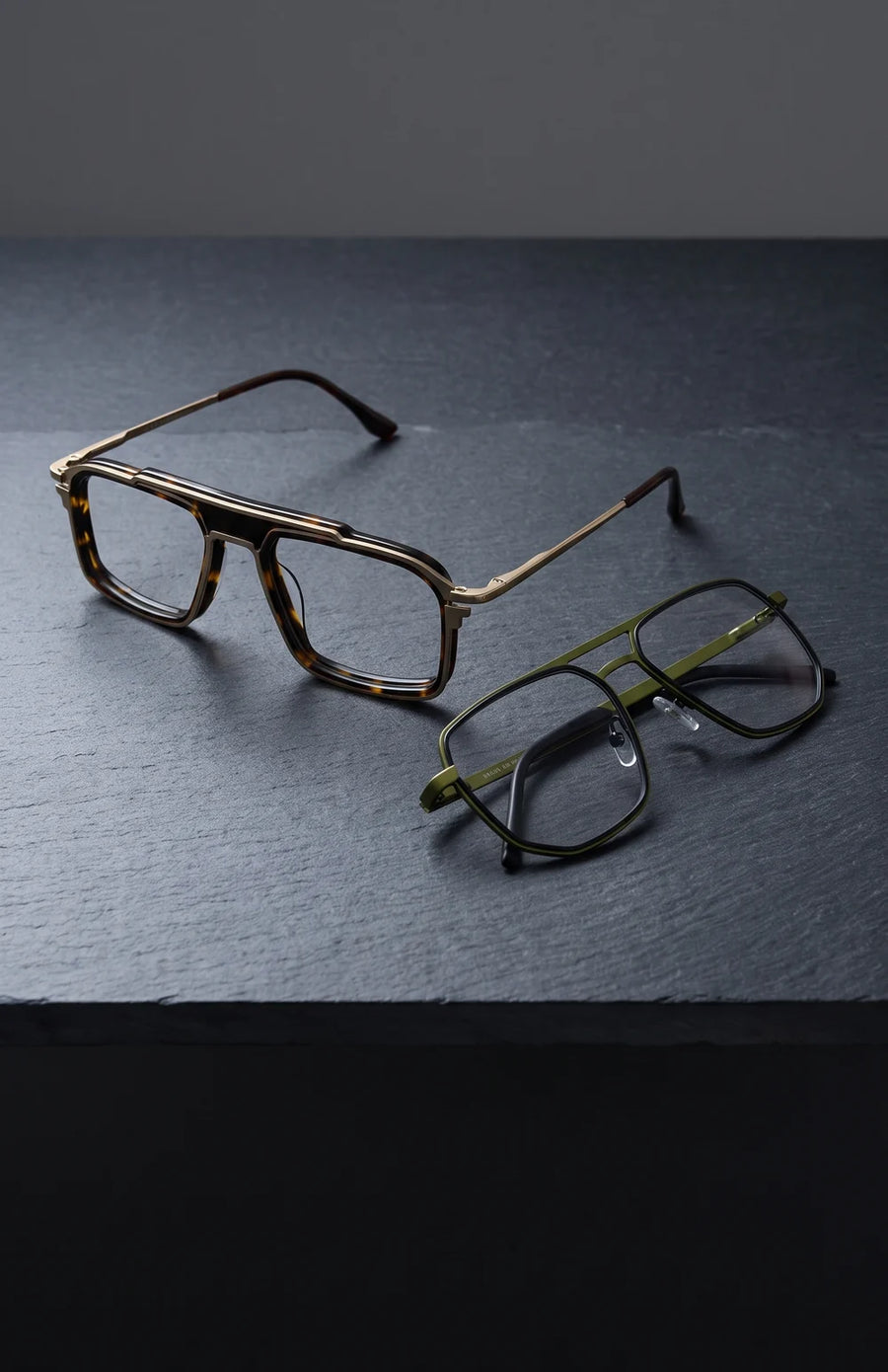 Two eyeglasses with double bridges, tortoise and gold, and green metal, on dark slate.