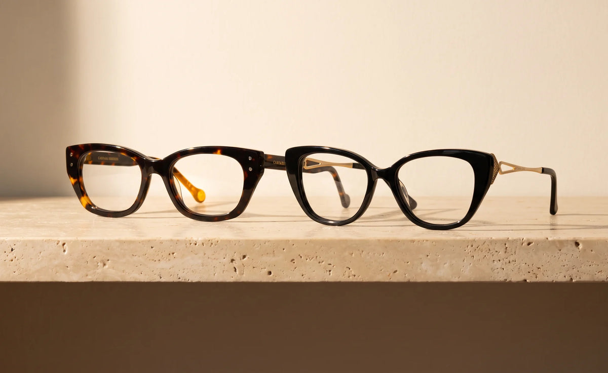 Thora in tortoise beside Vesper in black with a gold temple, two acetate eyeglasses on a cream travertine ledge in warm window light.