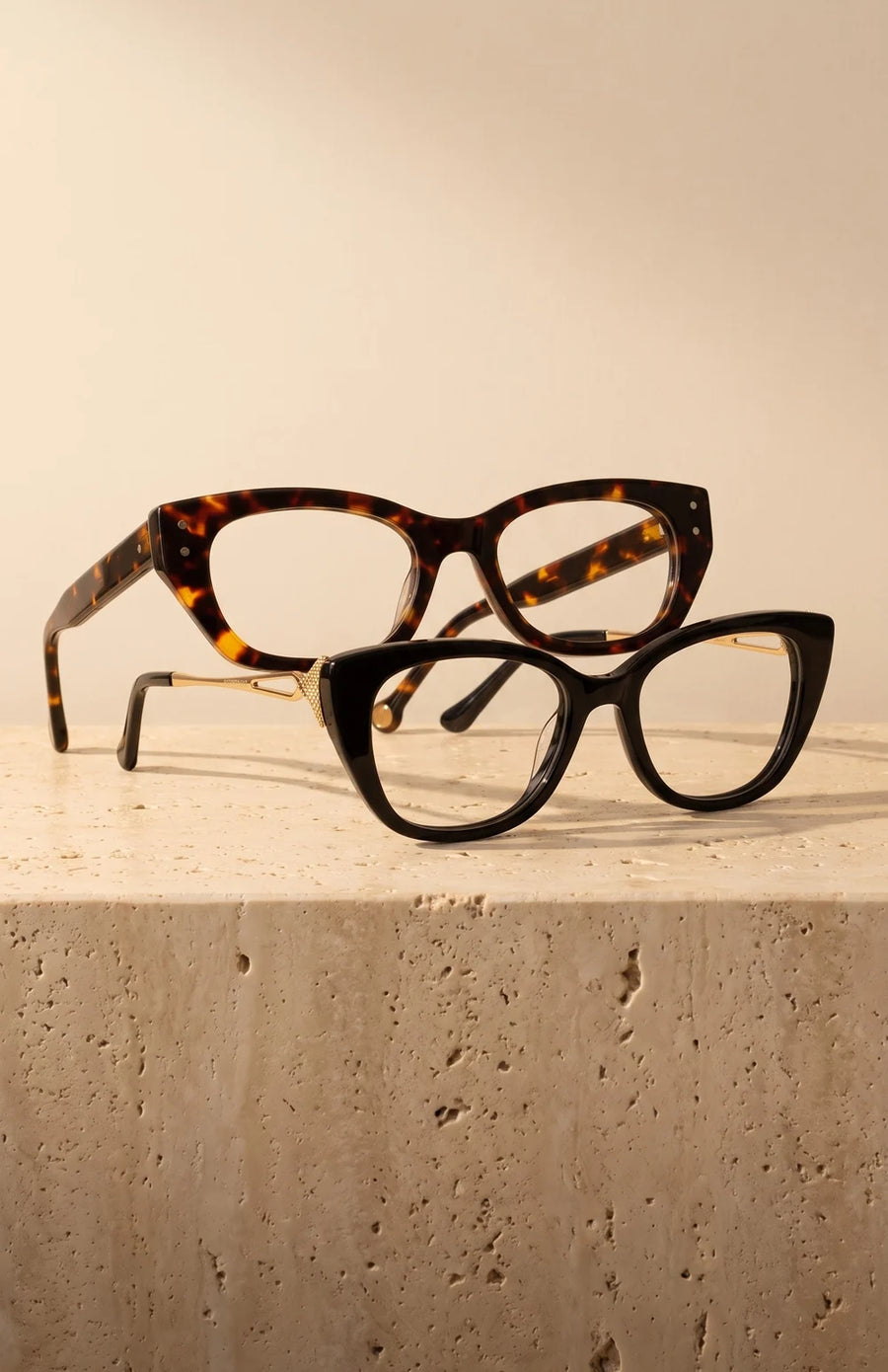 Two acetate eyeglasses, tortoise and black with a gold temple, on a cream stone ledge.