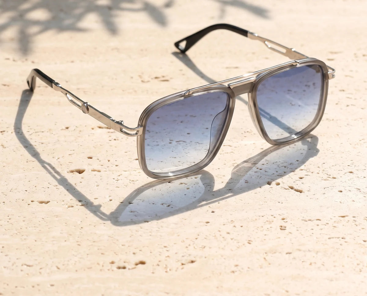 Cinder sunglasses in silver metal and smoke grey with blue gradient lenses on sunlit travertine.
